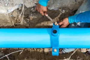 Person Tightening Bolts On A Blue Pipe With A Wrench In A Trench, Ensuring Seamless Water Line Repair.