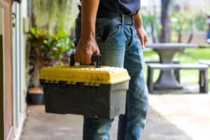 A Person Holds A Yellow And Black Toolbox Outdoors, Wearing Jeans And A Dark Shirt, Standing On A Patio With Lush Greenery In The Background—Ready For Efficient Debris Removal Service.