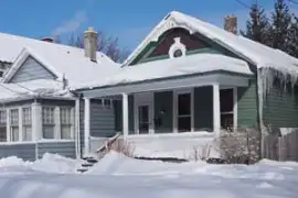 A Snow-Covered House With A Green Exterior And Front Porch Sits Alongside A Similar Home, Both Glistening In The Cold Weather. Icicles Hang From The Roof Under A Clear Blue Sky, Creating A Serene Winter Scene.
