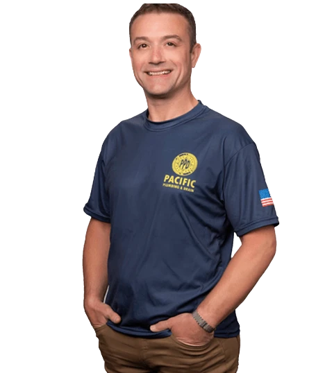 A man stands smiling with hands in pockets, wearing a navy blue Pacific Plumbing & Drain t-shirt and brown pants.