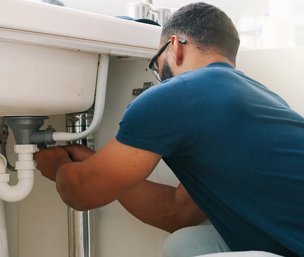 A person wearing glasses and a blue shirt is fixing plumbing pipes under a sink.
